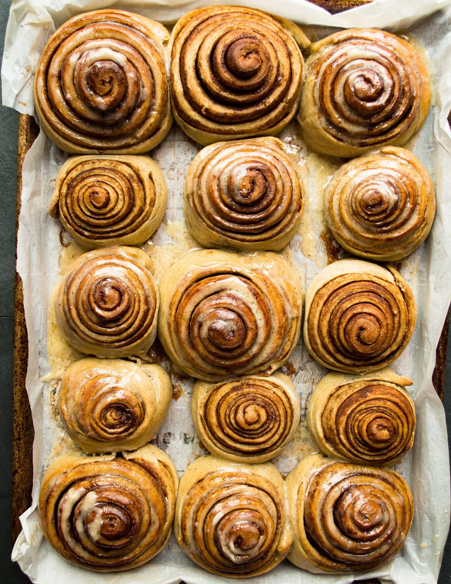 Overhead view of fifteen freshly baked Cinnamon Rolls with Honey Mascarpone Icing arranged in a 3-by-5 grid in a parchment-lined metal baking pan