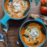 Overhead view of two bright teal crocks of Roasted Tomato and Red Pepper Bisque with Burrata and Garlic Tostada on a warm wooden board