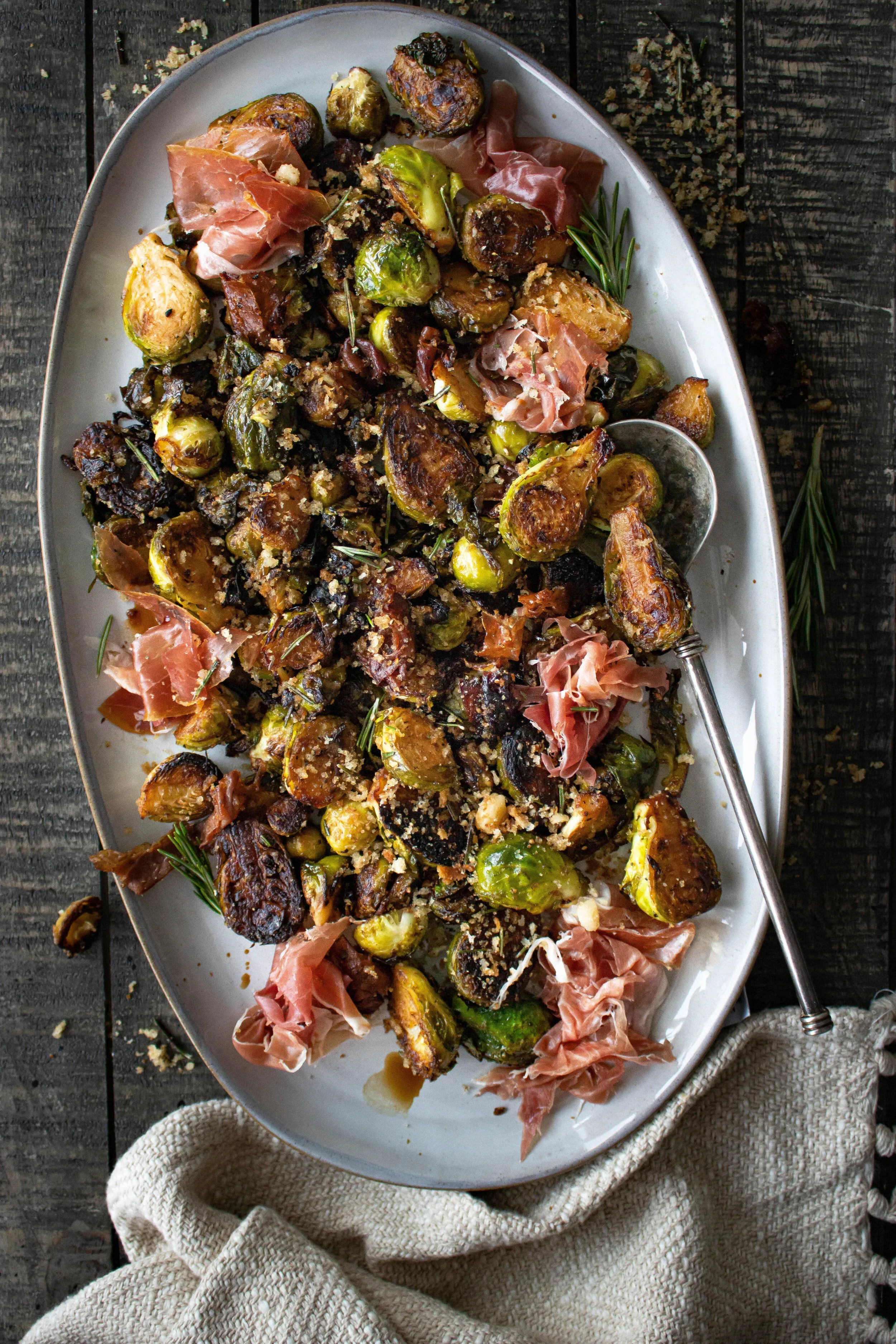 Pan Fried Brussels Sprouts with prosciutto and herby breadcrumbs mounded on a large white oval ceramic platter on dark weathered wood