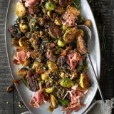 Pan Fried Brussels Sprouts with prosciutto and herby breadcrumbs mounded on a large white oval ceramic platter on dark weathered wood