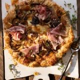 Whole Mushroom and Caramelized Onion Phyllo Tart with Prosciutto resting on a white handled cutting board over a slatted wood surface
