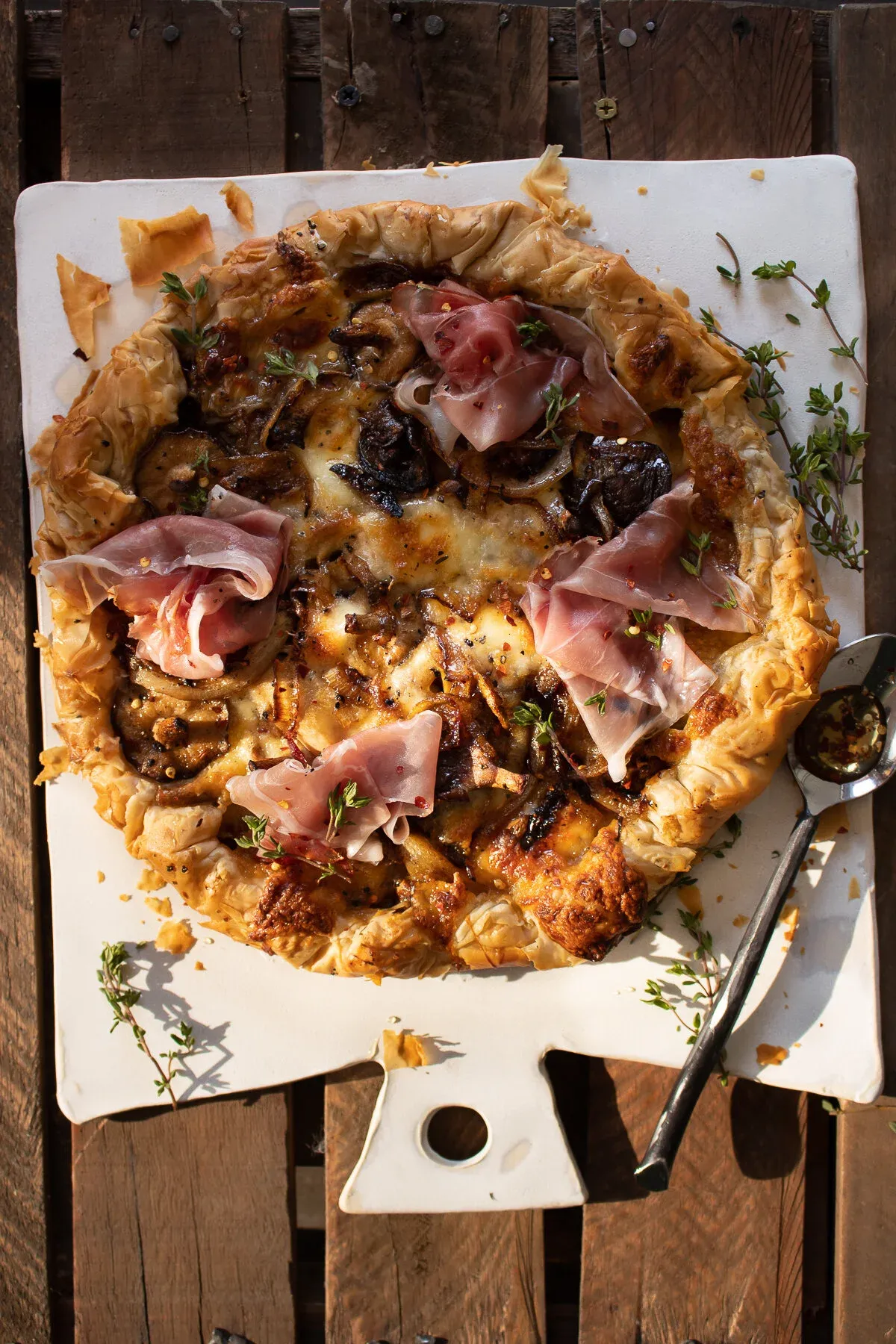 Whole Mushroom and Caramelized Onion Phyllo Tart with Prosciutto resting on a white handled cutting board over a slatted wood surface