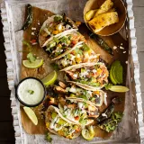 Overhead flat-lay of five Blackened Salmon Tacos with Grilled Pineapple and Mango Salsa arranged on kraft paper inside a carved whitewashed wood tray