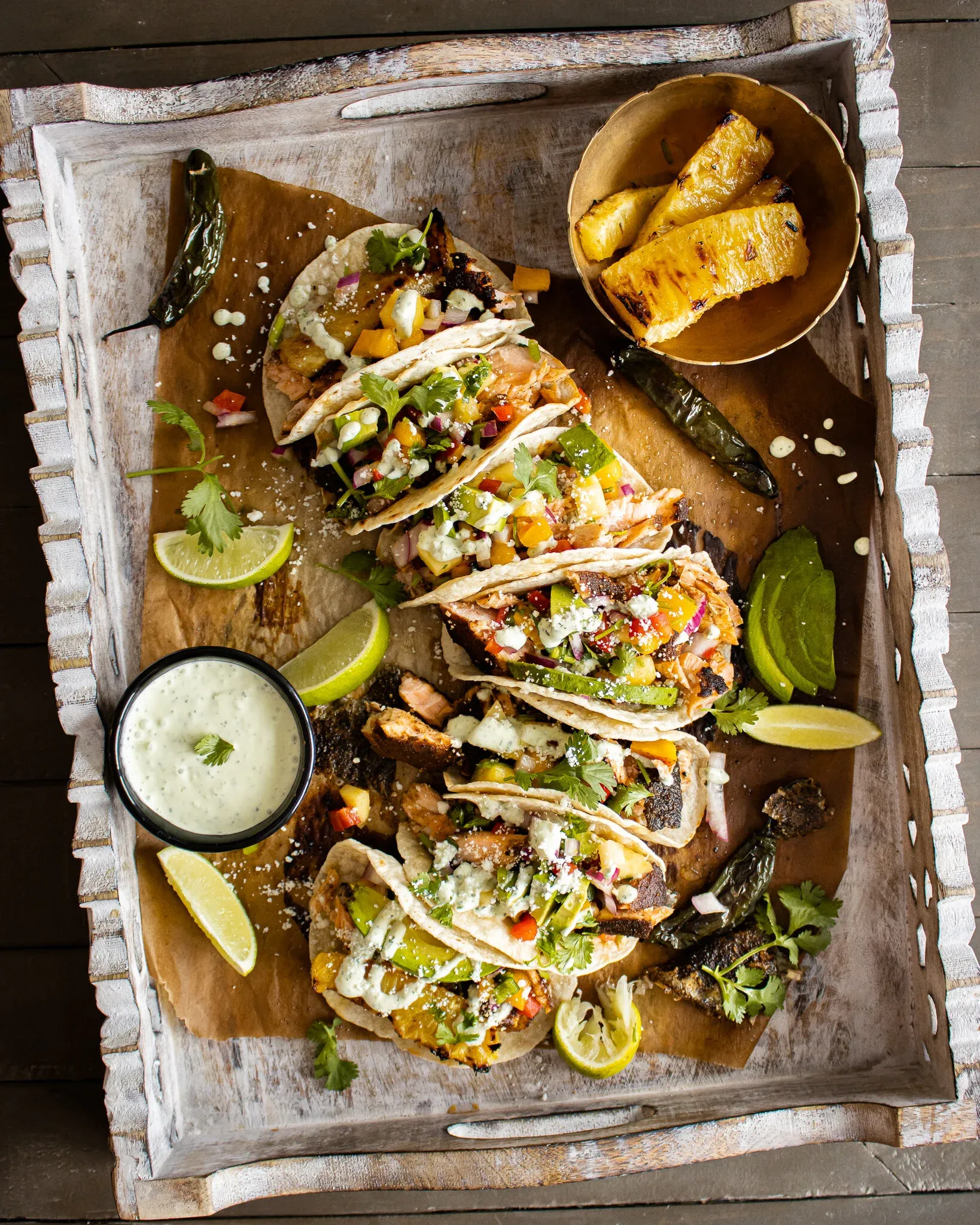 Overhead flat-lay of five Blackened Salmon Tacos with Grilled Pineapple and Mango Salsa arranged on kraft paper inside a carved whitewashed wood tray