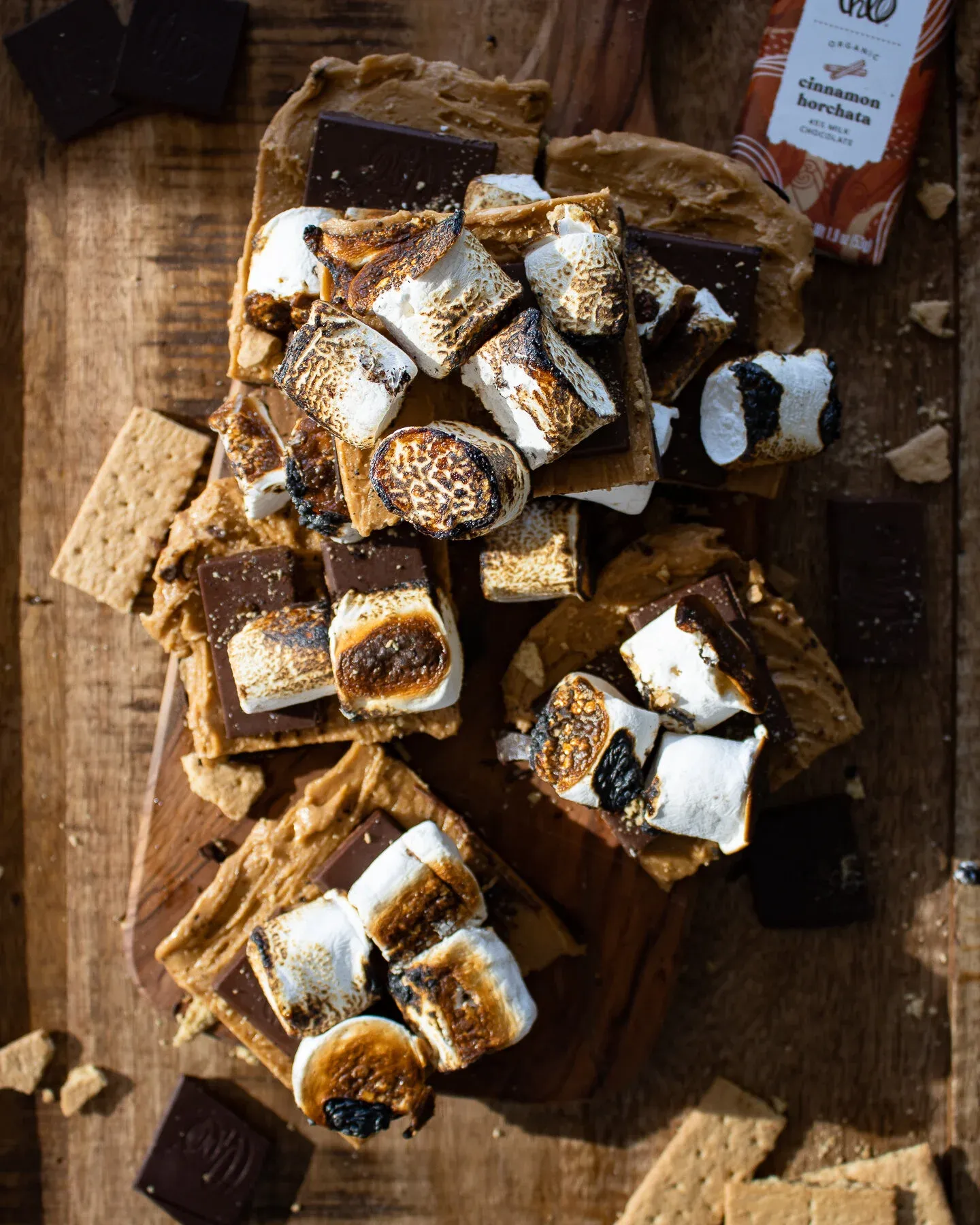 Overhead shot of cut No Bake Peanut Butter and Horchata S'mores Bars piled on a small wooden cutting board on a dark wood surface