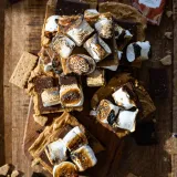 Overhead shot of cut No Bake Peanut Butter and Horchata S'mores Bars piled on a small wooden cutting board on a dark wood surface