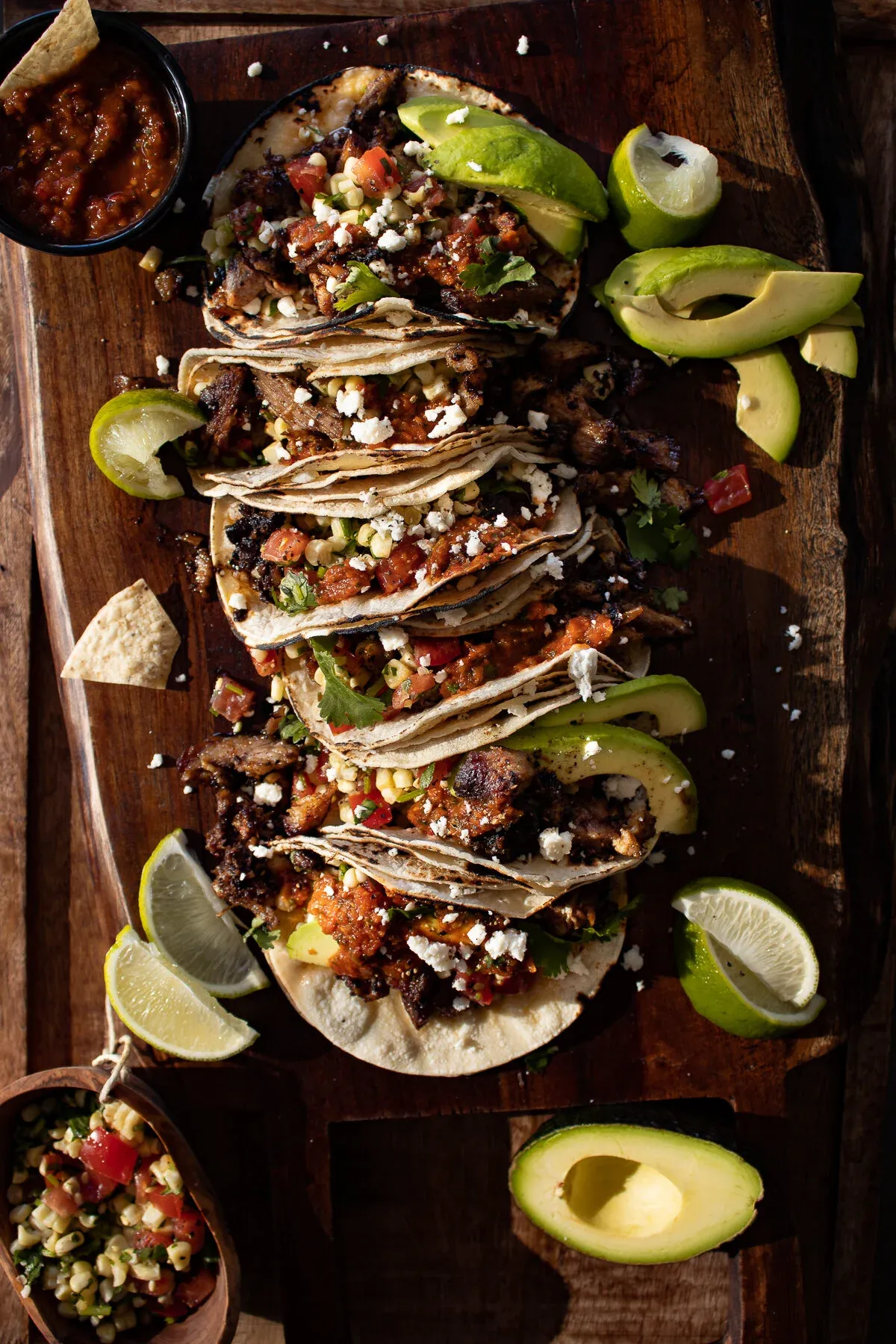 Pernil Tacos lined up diagonally on a large dark wooden cutting board