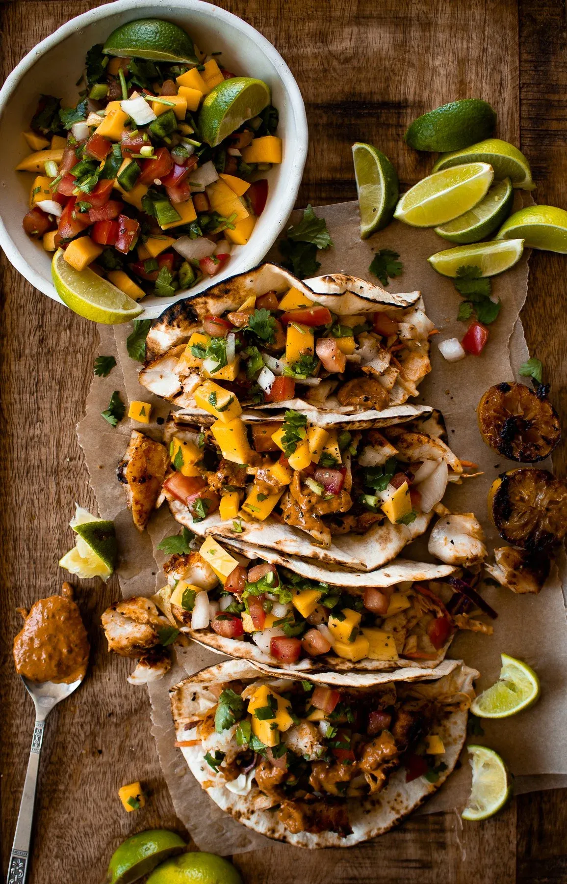 Overhead flat-lay of four Caribbean fish tacos with mango salsa lined up on crinkled parchment paper on a wooden cutting board