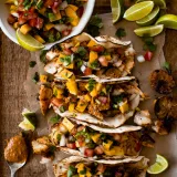 Overhead flat-lay of four Caribbean fish tacos with mango salsa lined up on crinkled parchment paper on a wooden cutting board