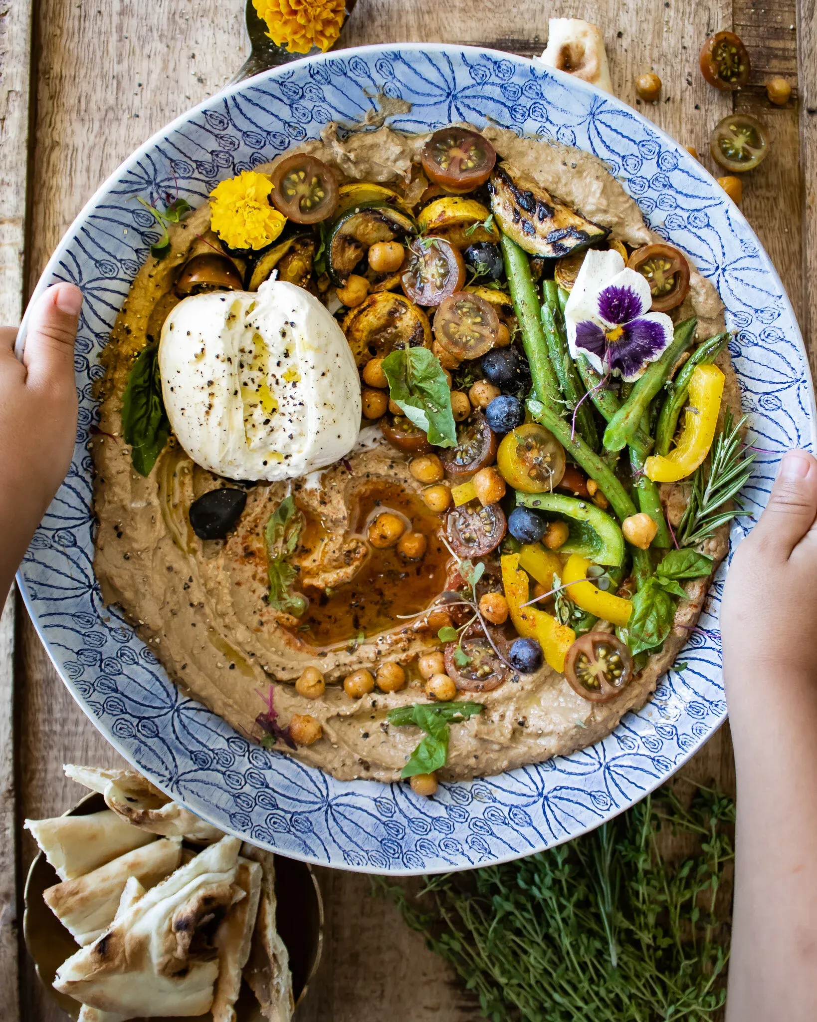 Overhead view of a Black Garlic Hummus Platter with Summer Veggies and Burrata served in a large blue-and-white patterned ceramic plate held by two hands