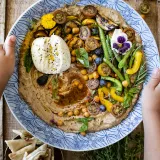 Overhead view of a Black Garlic Hummus Platter with Summer Veggies and Burrata served in a large blue-and-white patterned ceramic plate held by two hands