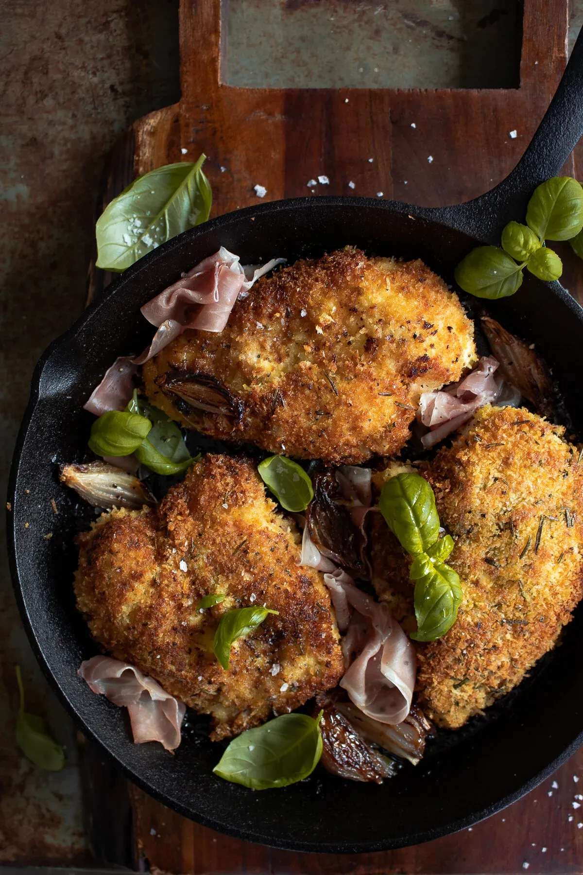 Three golden panko-crusted Prosciutto and Basil Wrapped Chicken breasts in a black cast iron skillet on a wood cutting board