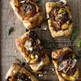 Five caramelized leek and peach puff pastry tarts scattered casually on a rustic wooden cutting board