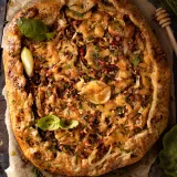 Overhead view of a whole cheesy potato, onion and chive tart with pancetta on parchment paper on a dark metal baking sheet