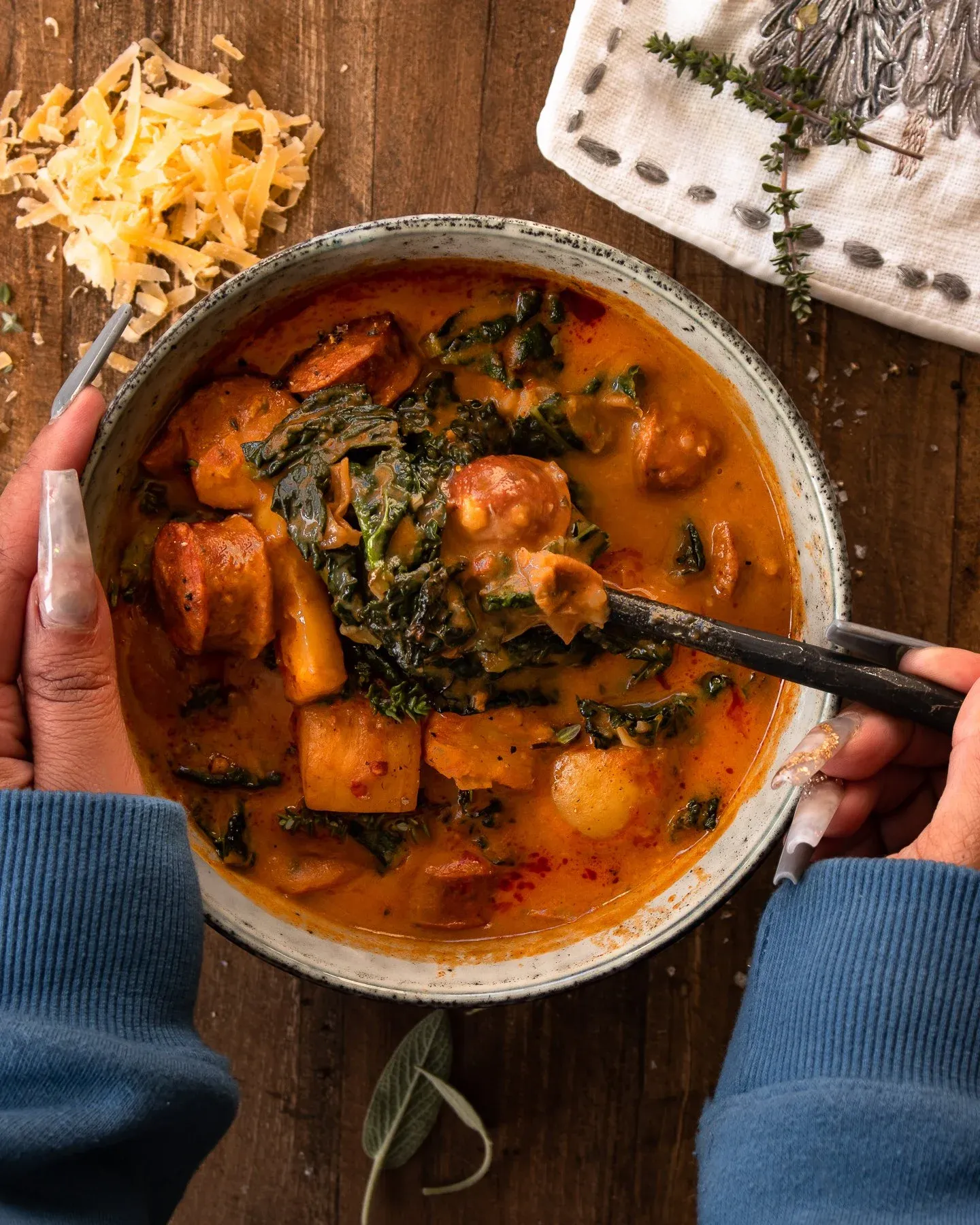 Smoked Potato and Sausage Soup in a speckled grey ceramic bowl held by two hands in blue sleeves, overhead view on dark wood