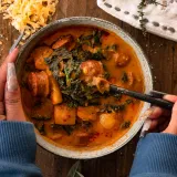 Smoked Potato and Sausage Soup in a speckled grey ceramic bowl held by two hands in blue sleeves, overhead view on dark wood
