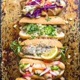 Five Better Than Basic Sausage Dogs lined up on a dark seasoned sheet pan, each with a different gourmet topping