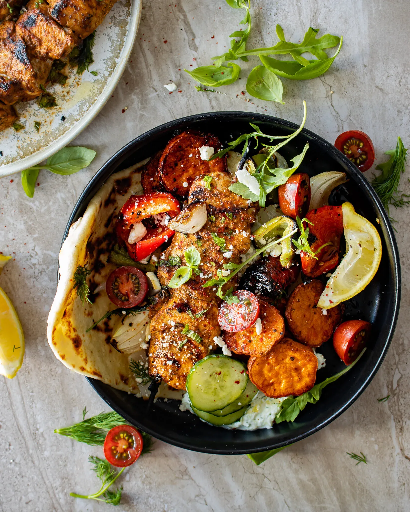 Overhead view of a black ceramic bowl with a serving of chicken and vegetable skewers with roasted sweet potatoes