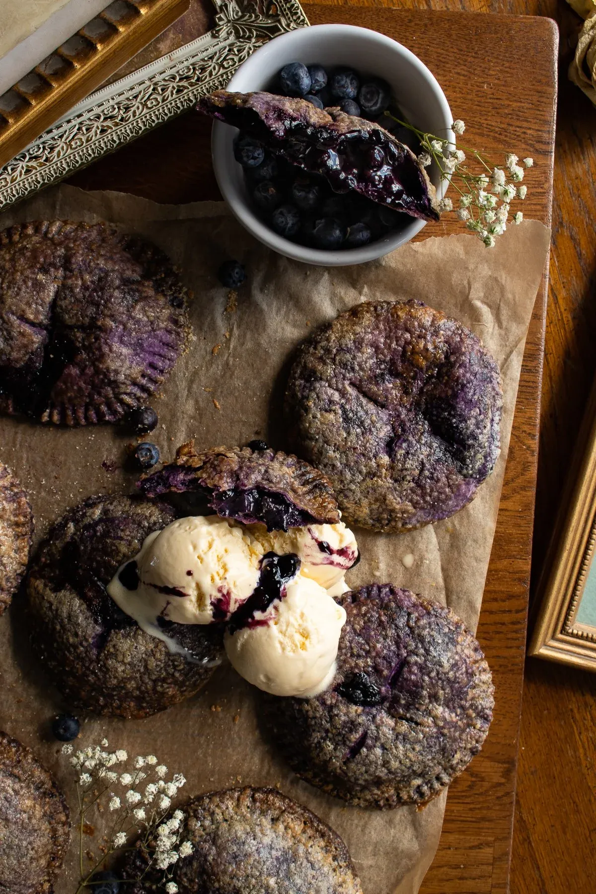 Multiple Ube and Blueberry Mini Pies on parchment paper on a dark wooden board, angled overhead view