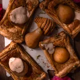 Overhead dark-moody flat-lay of five rectangular poached pear pastry tarts with mascarpone cream arranged on a white ceramic surface atop a dark wood table