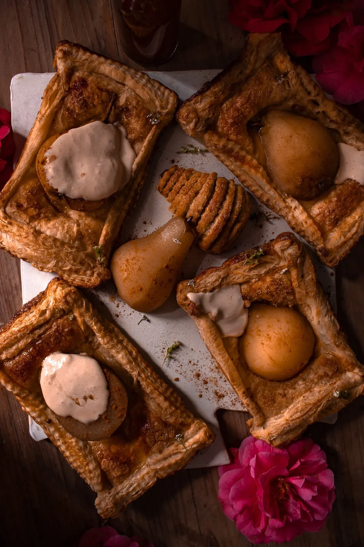 Overhead dark-moody flat-lay of five rectangular poached pear pastry tarts with mascarpone cream arranged on a white ceramic surface atop a dark wood table