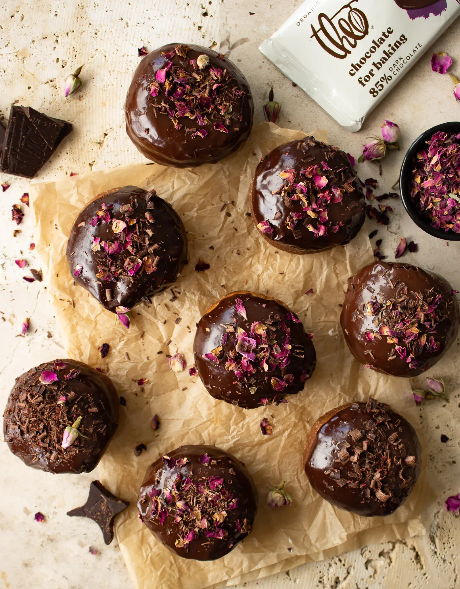Nine cardamom and rose brioche doughnuts with dark chocolate glaze arranged on crinkled parchment paper on a cream surface