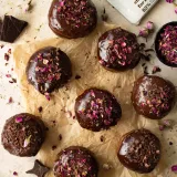 Nine cardamom and rose brioche doughnuts with dark chocolate glaze arranged on crinkled parchment paper on a cream surface