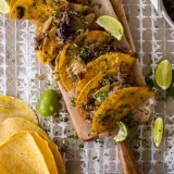 Eight Crispy Dominican Beef Tacos lined up on a long narrow wooden paddle board over parchment paper