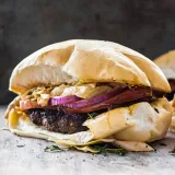 Dominican Chimi Burger served on a soft white pan de agua roll stuffed with a thick charred beef patty, sliced ripe tomato