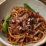 White rustic bowl of Dominican Style Goat Ragù served over thick bucatini pasta coated in a deep-red tomato-braised goat sauce