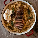 Overhead shot of Brown Butter Orzo with Steak in a white enamel wide pan with red handles; toasted amber orzo fills the pan
